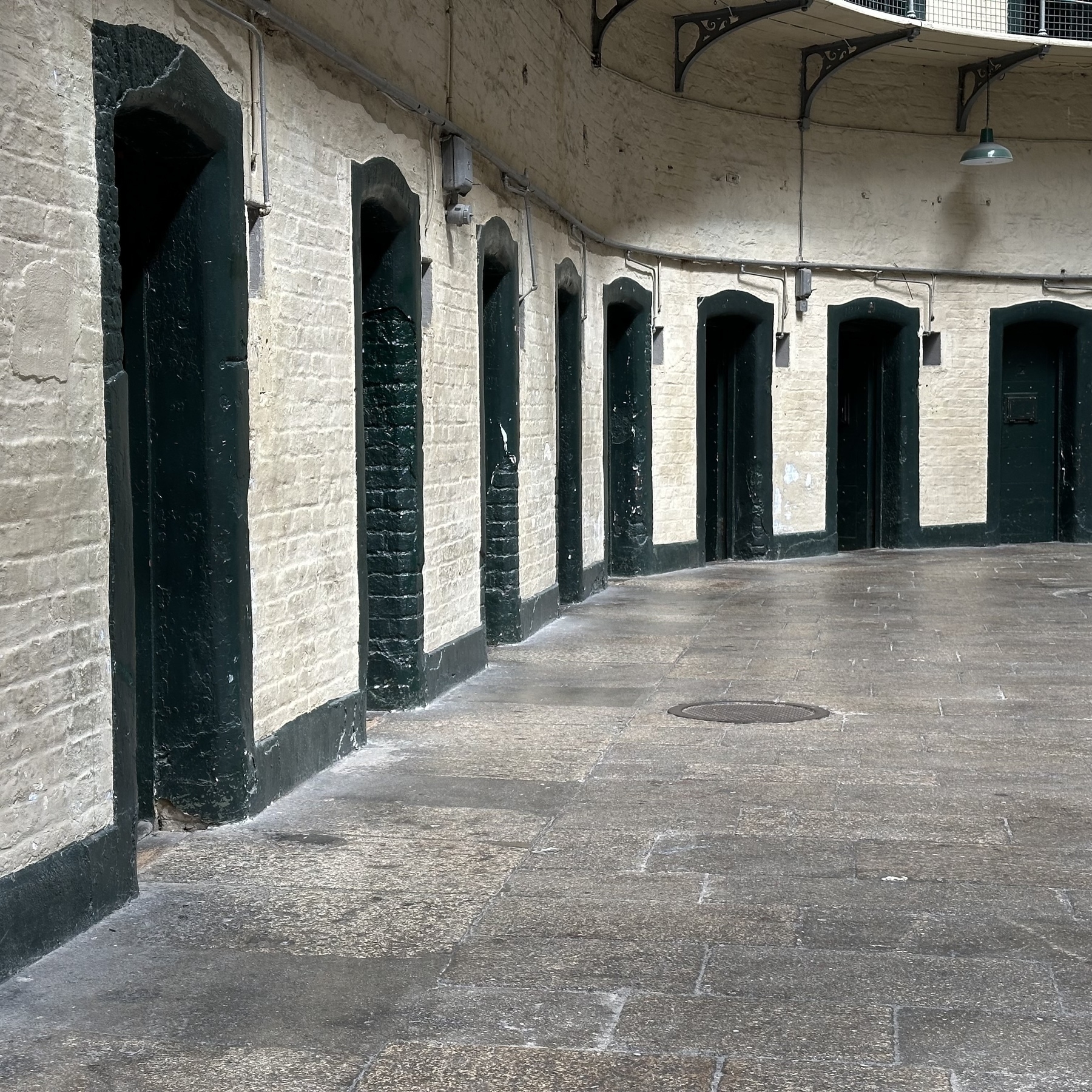 Auto-generated description: A narrow, enclosed corridor features multiple dark green doors set into aged brick walls with a paved stone floor.