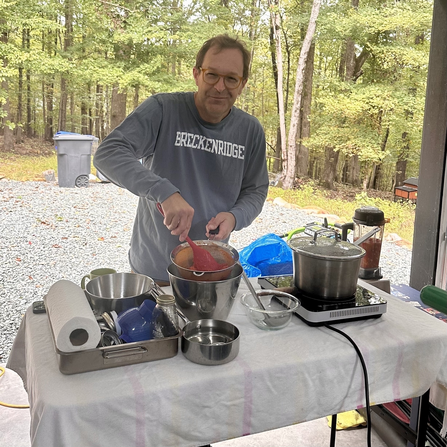 Anton is cooking outdoors at a table with various kitchen supplies and appliances, set against a backdrop of trees and a gravel path.